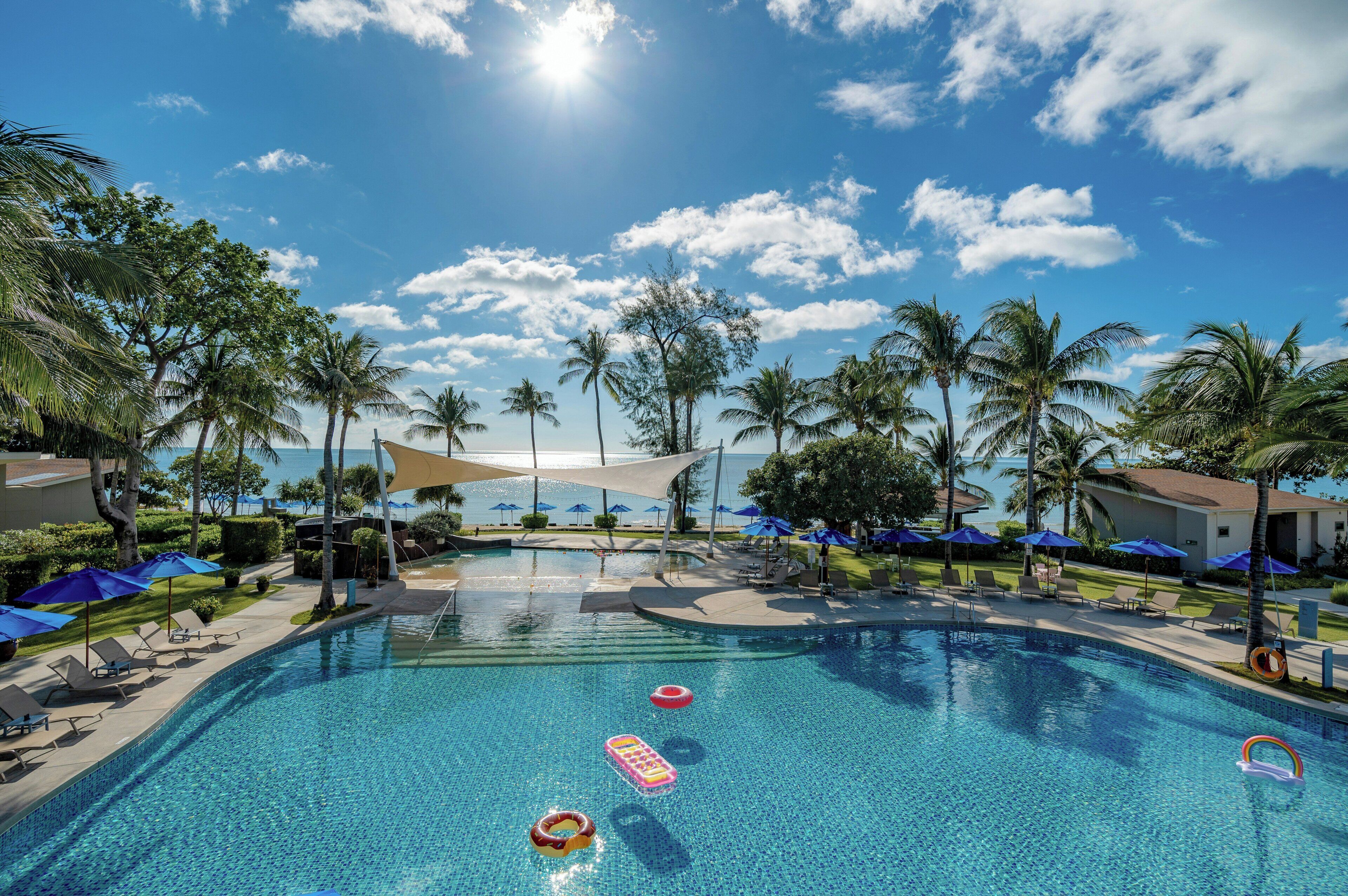 A swimming pool with lounge chairs and umbrellas, surrounded by palm trees and a clear sky.