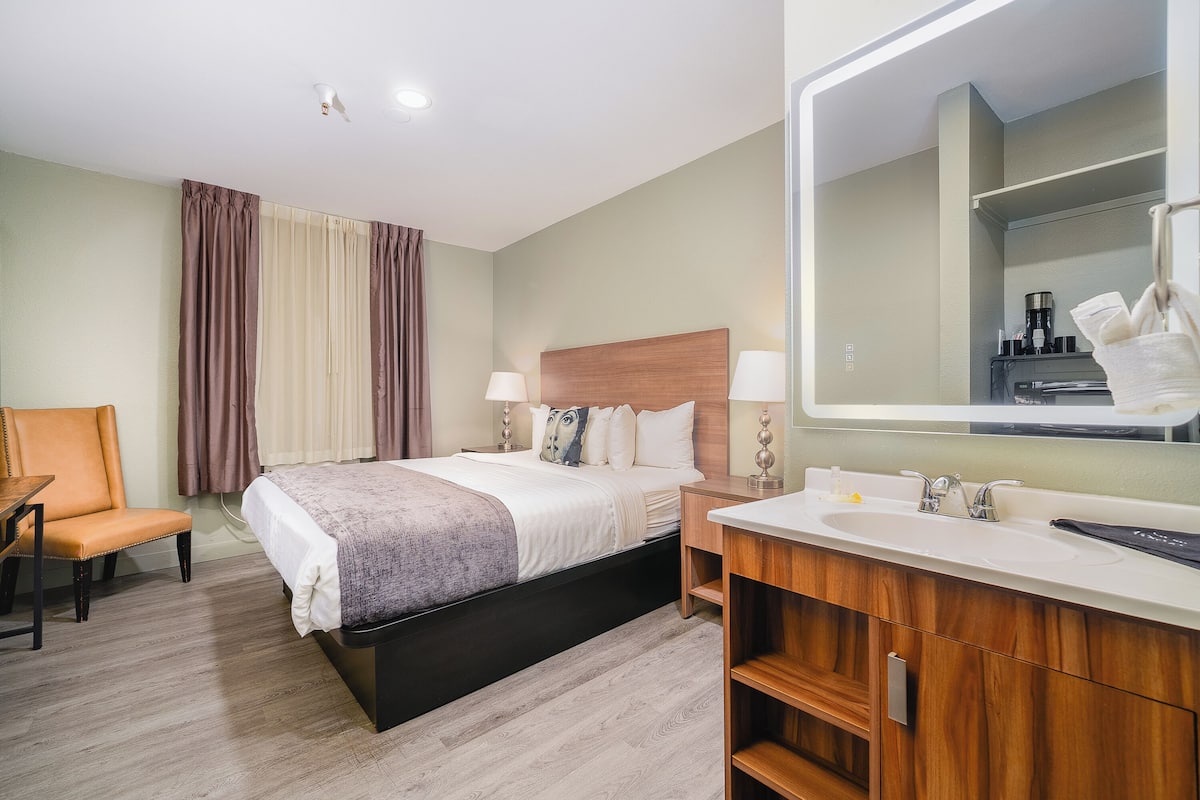 Une chambre d’hôtel comprenant un lit, une chaise, une table de chevet, un miroir et une salle de bain avec un lavabo et un miroir.