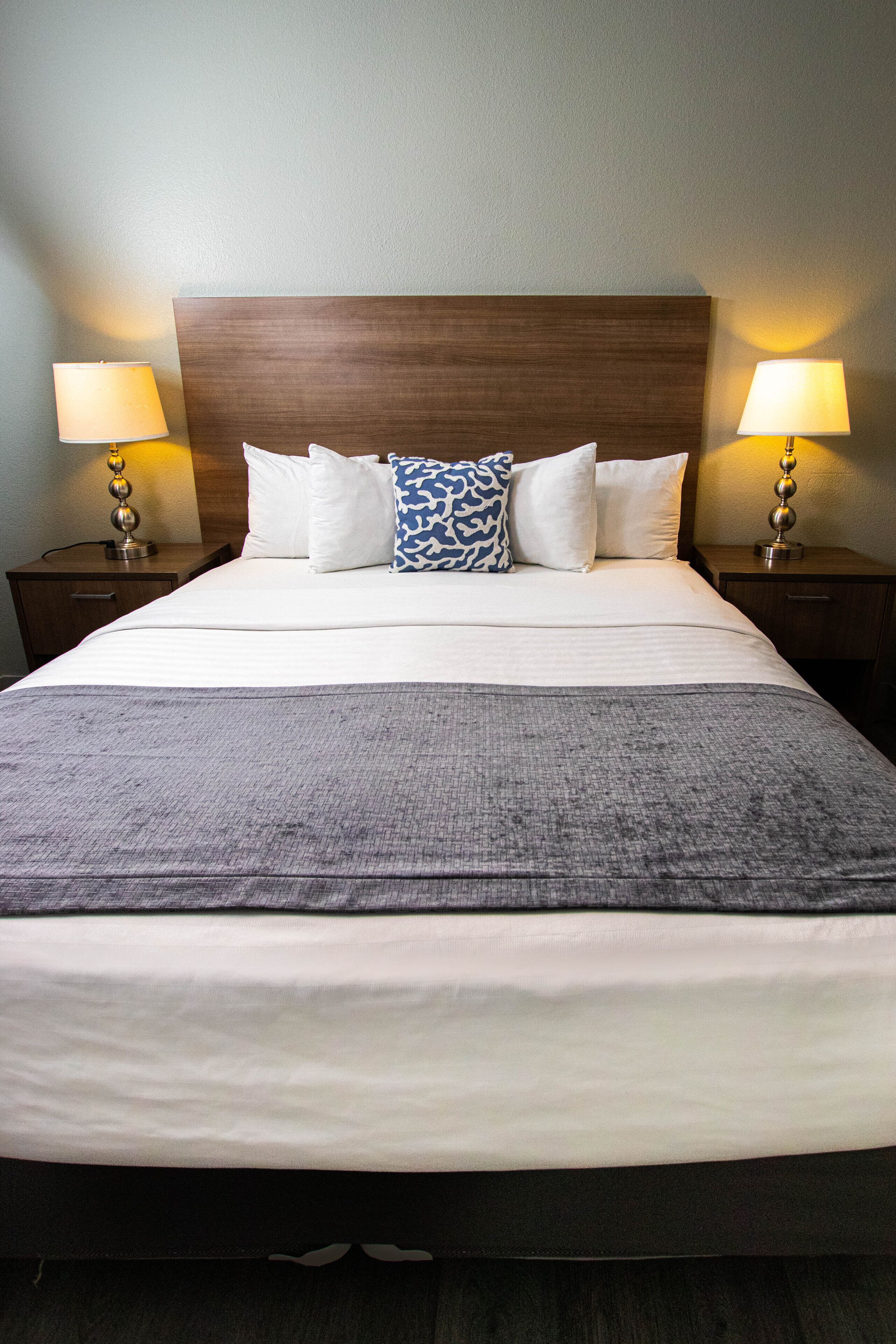 Ein ordentlich gemachtes Bett mit weißen und blauen Kissen, einer grauen Tagesdecke und zwei Nachttischlampen auf Holznachttischen.