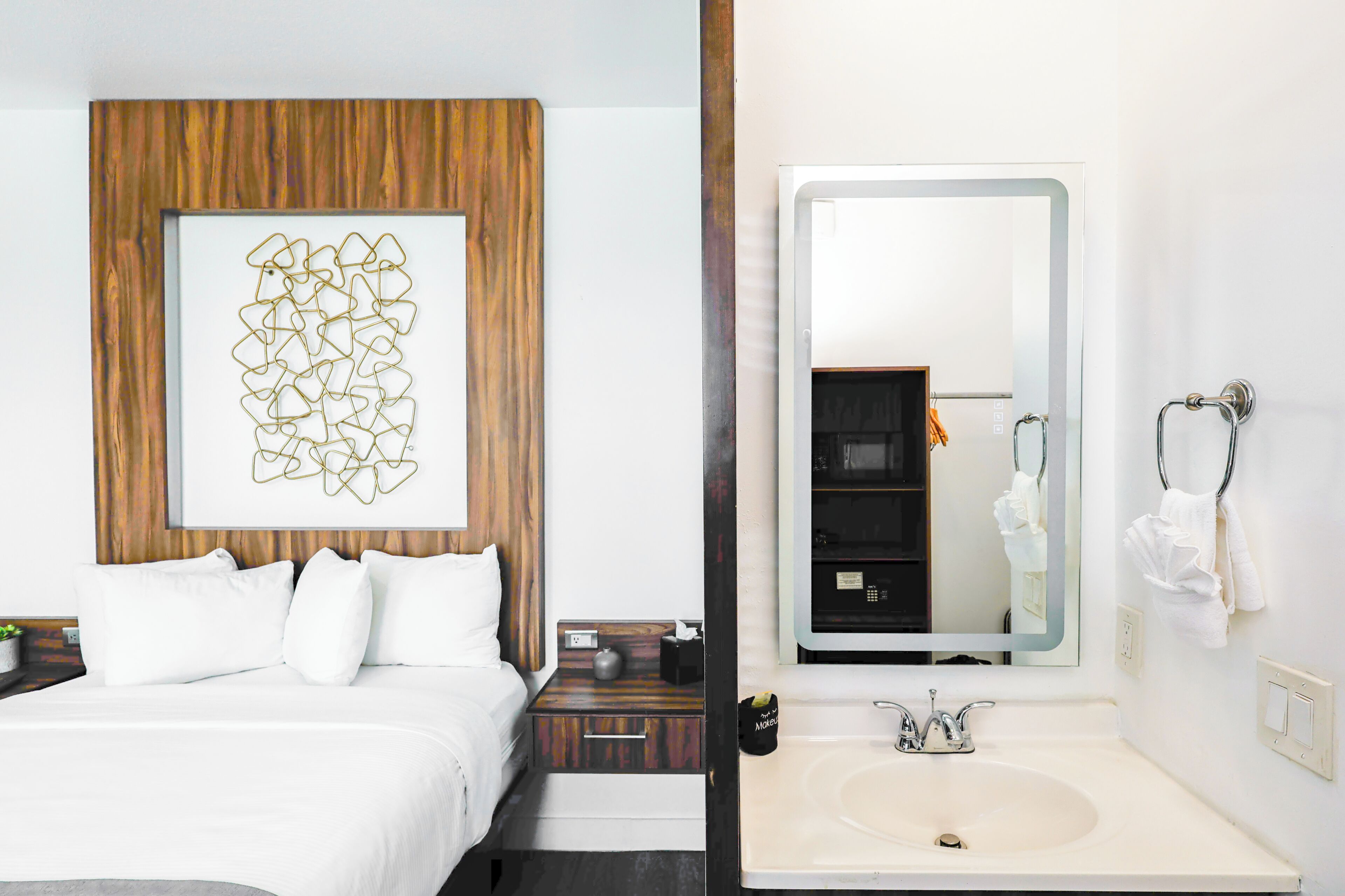 Quarto de hotel com uma cama, uma obra de arte na parede, uma mesa de cabeceira e um banheiro com pia e espelho.