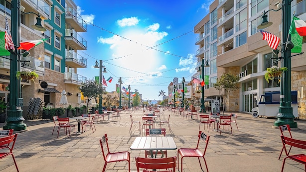 Un café al aire libre con sillas rojas y mesas blancas, rodeado de edificios con balcones y banderas.