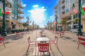 Un café al aire libre con sillas rojas y mesas blancas, rodeado de edificios con balcones y banderas.