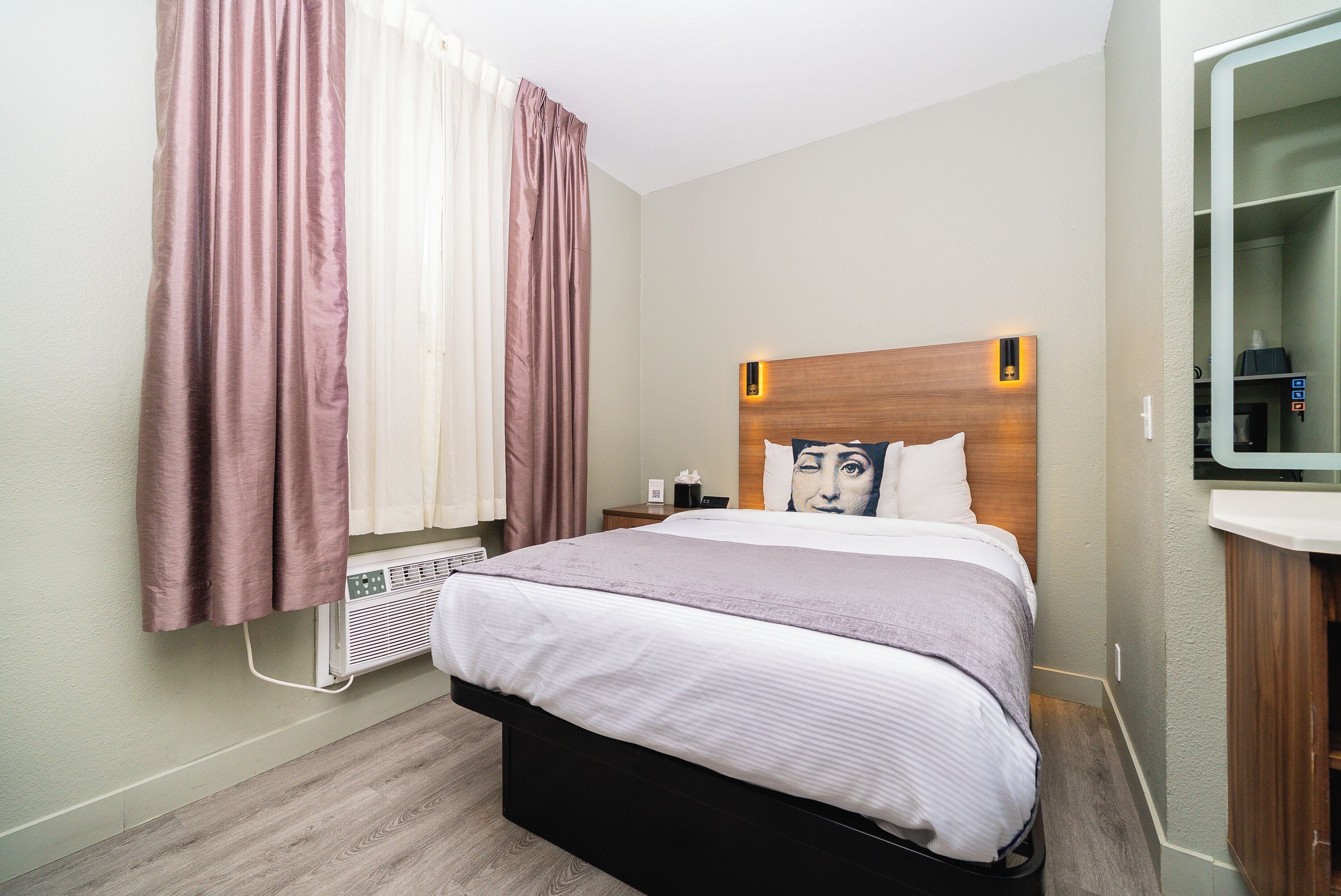 A hotel room with a bed, a bedside table, a mirror, and a window with curtains.