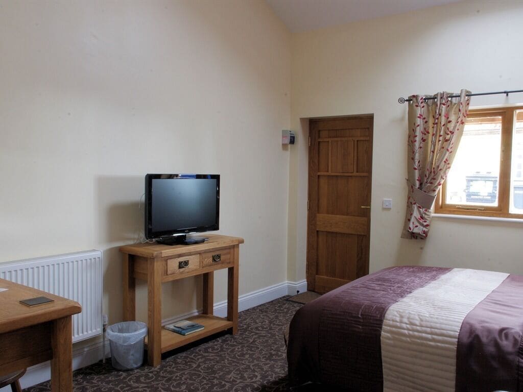 A hotel room with a bed, a TV on a wooden stand, a door, and a window with curtains.