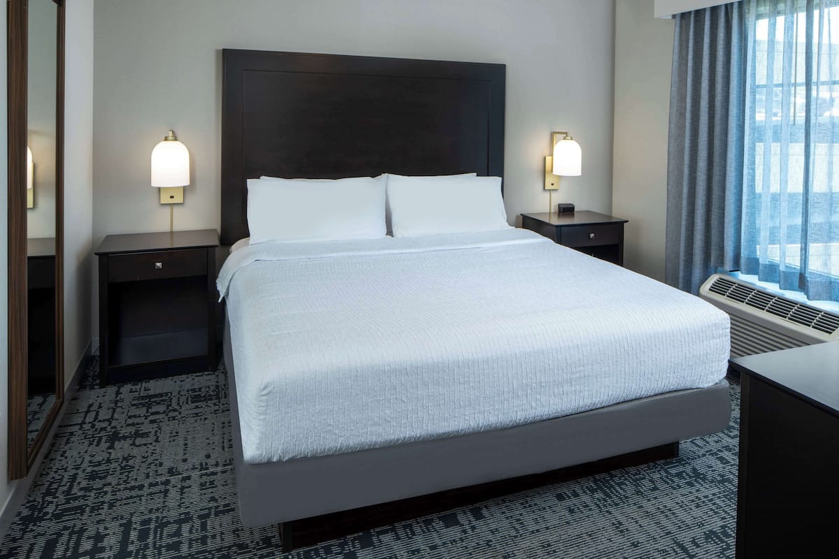 A hotel room with a large bed, two bedside tables with lamps, a dark headboard, and a view of the city through large windows.