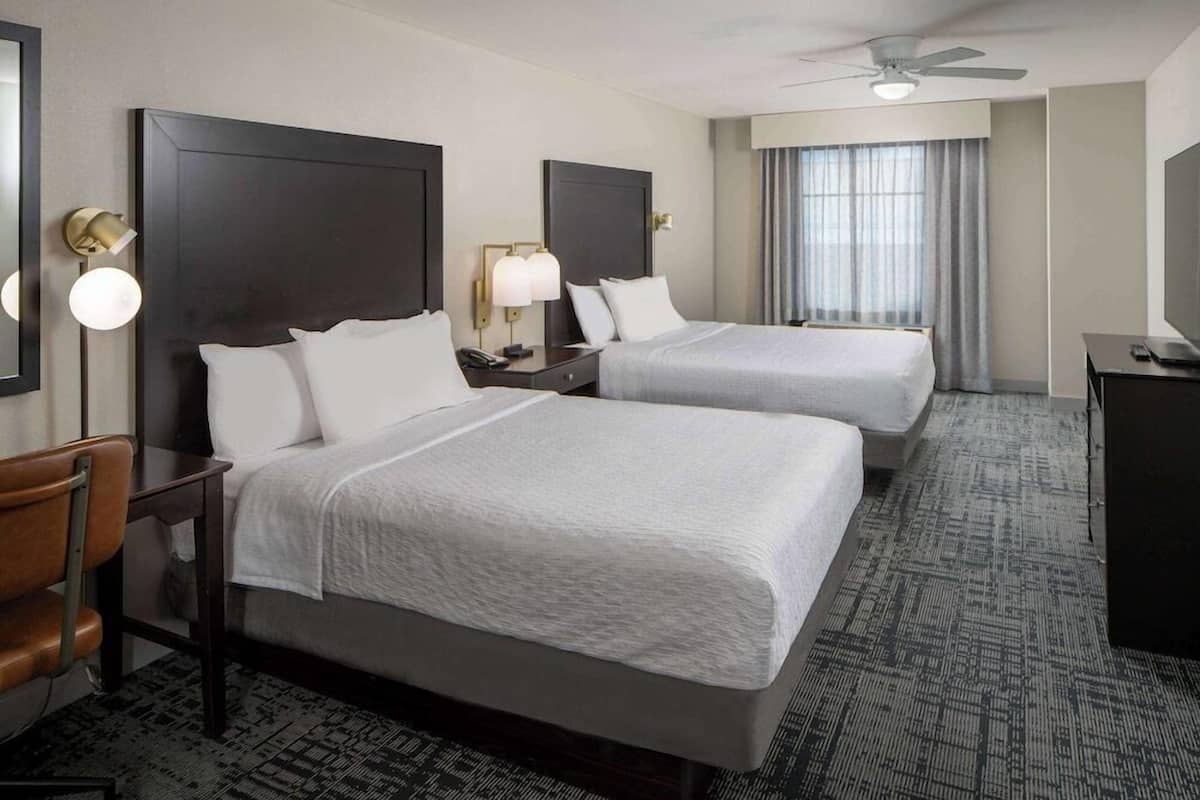 A hotel room with two beds, a desk, a chair, a TV, and a ceiling fan.