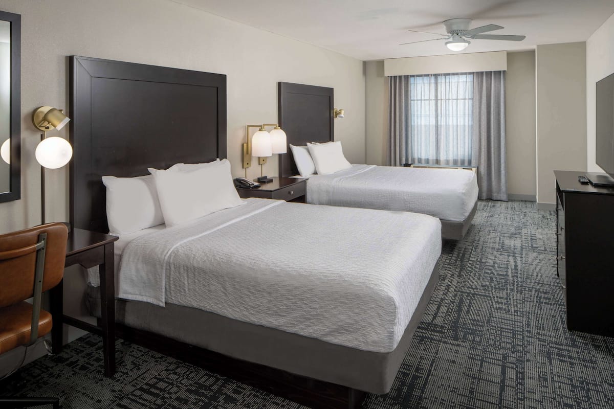 A hotel room with two beds, a desk, a chair, a TV, and a ceiling fan.