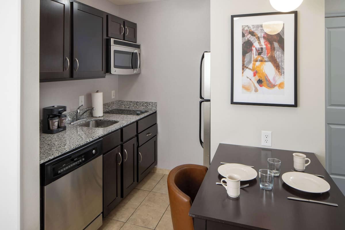 A compact kitchen with dark cabinets, a microwave, a dishwasher, and a dining table set with plates, cups, and a glass.