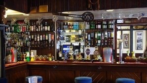 A bar area with wooden paneling, a variety of liquor bottles, a cash register, and two stools. - The Bugle Inn (St Austell)