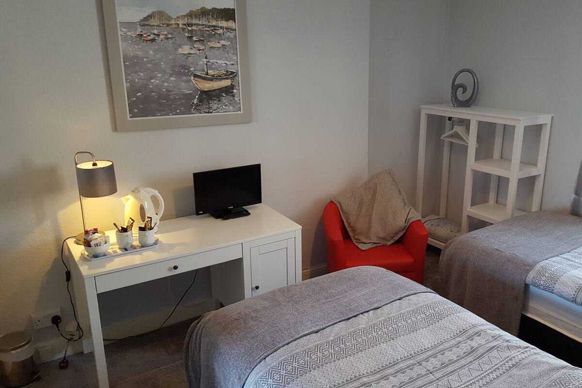 A hotel room with a desk, a television, a red chair, two beds, and a painting of boats on the wall.