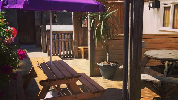 An outdoor wooden deck with picnic tables and a purple umbrella labeled "TRIBUTE CORNHISH PALE ALE". - The Bugle Inn (St Austell)