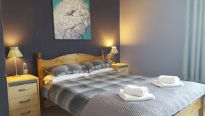 A neatly made bed with a plaid bedspread, two pillows, and two folded towels on a wooden headboard. There are two bedside tables with lamps and a water bottle on one. A large floral painting hangs on the wall above the bed. - The Bugle Inn (St Austell)