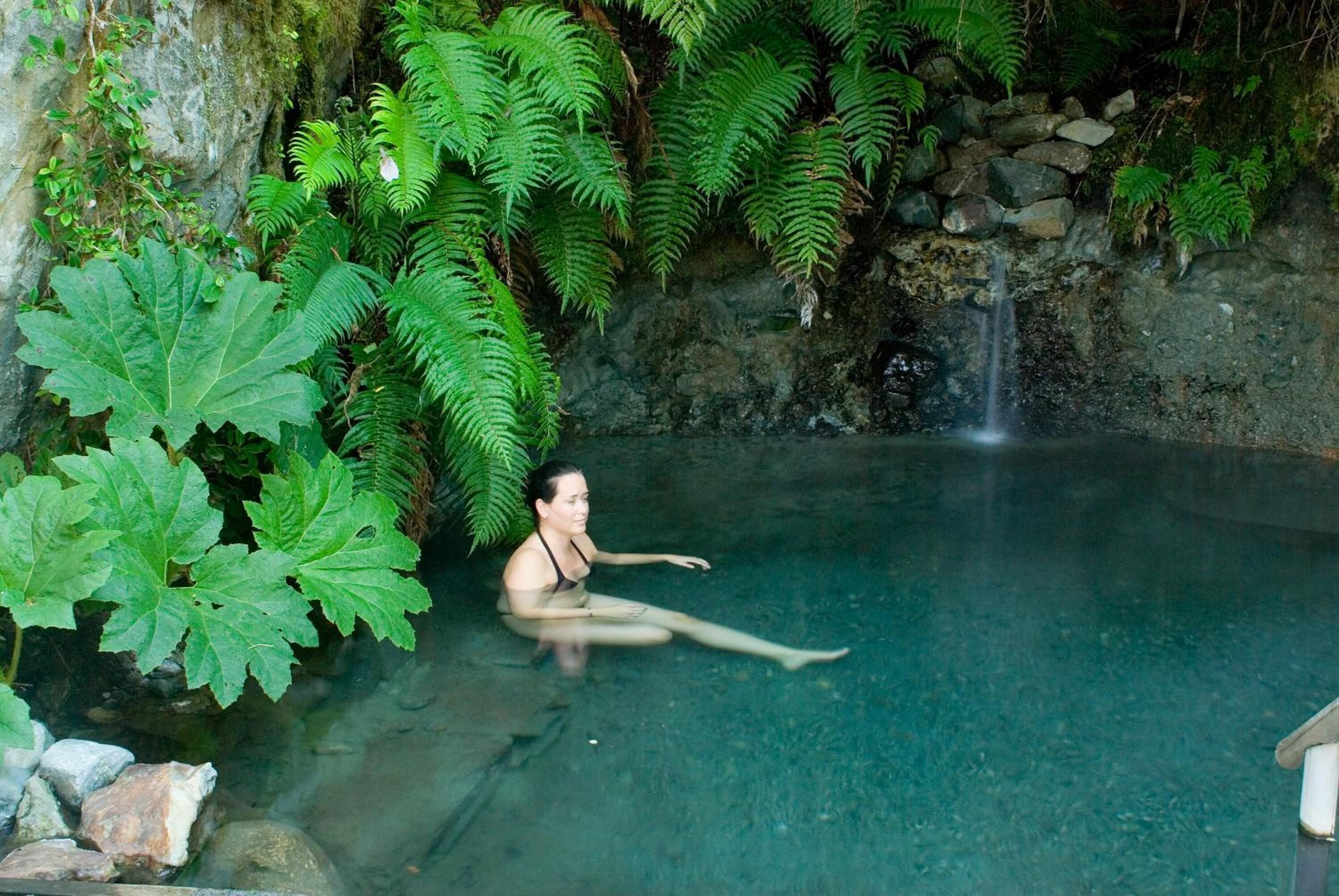 Una dona asseguda en una piscina transparent rodejada de vegetació exuberant i una petita cascada.