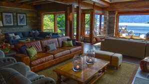 A spacious living room with a large brown leather sofa, a beige sofa, and a wooden coffee table with candles. There are framed pictures on the wall and a view of the ocean through large windows. - Puyuhuapi Lodge & Spa (Cisnes)