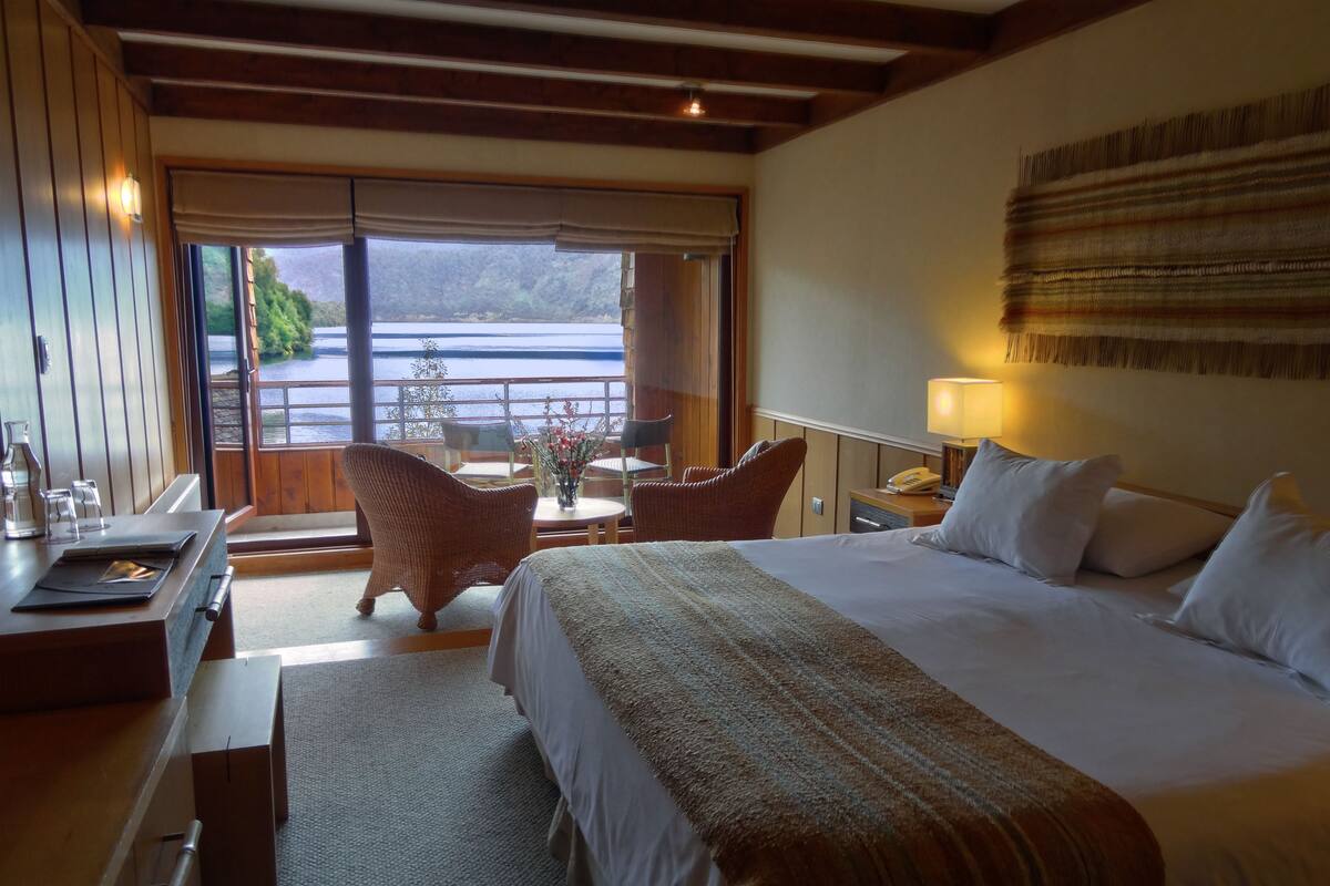 A hotel room with a large bed, a desk with a chair, a small table with a lamp, and a view of a lake through a window.