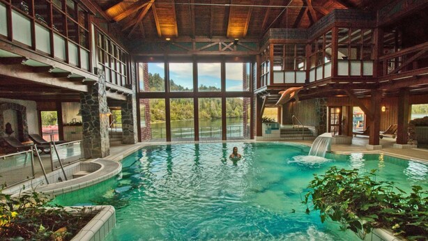 An indoor swimming pool with a waterfall feature, surrounded by stone walls and large windows offering scenic views. - Puyuhuapi Lodge & Spa (Cisnes)