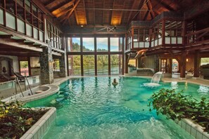An indoor swimming pool with a waterfall feature, surrounded by stone walls and large windows offering scenic views.