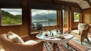 A room with a large window offering a view of mountains and a lake. There are wicker chairs and a table with a glass of drink. - Puyuhuapi Lodge & Spa (Cisnes)