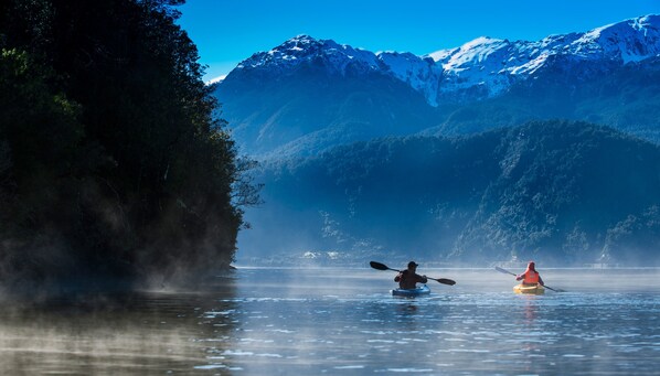 A calm lake with kayakers, surrounded by forested hills and snow-capped mountains.