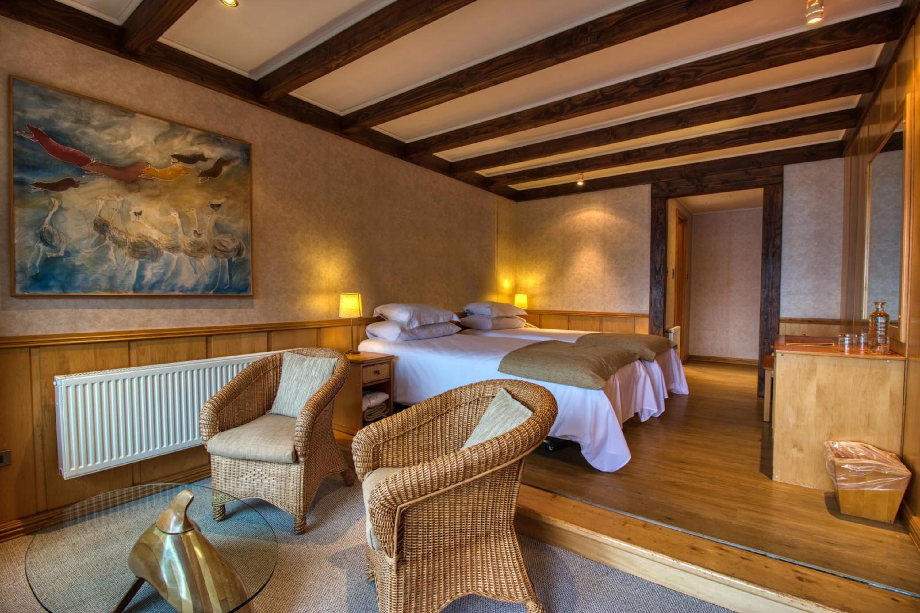 A cozy room with a bed, two wicker chairs, a radiator, and a painting on the wall.