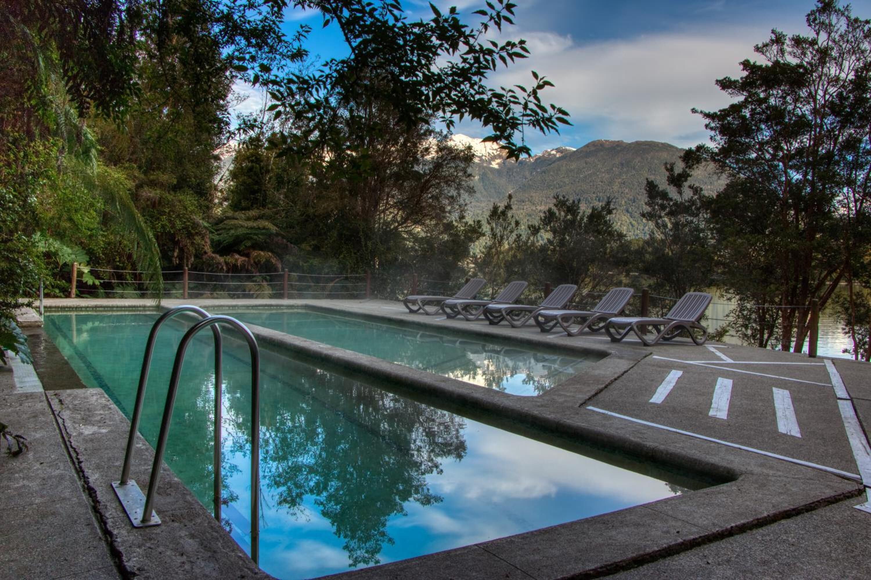 A swimming pool with lounge chairs, surrounded by trees and mountains in the background.