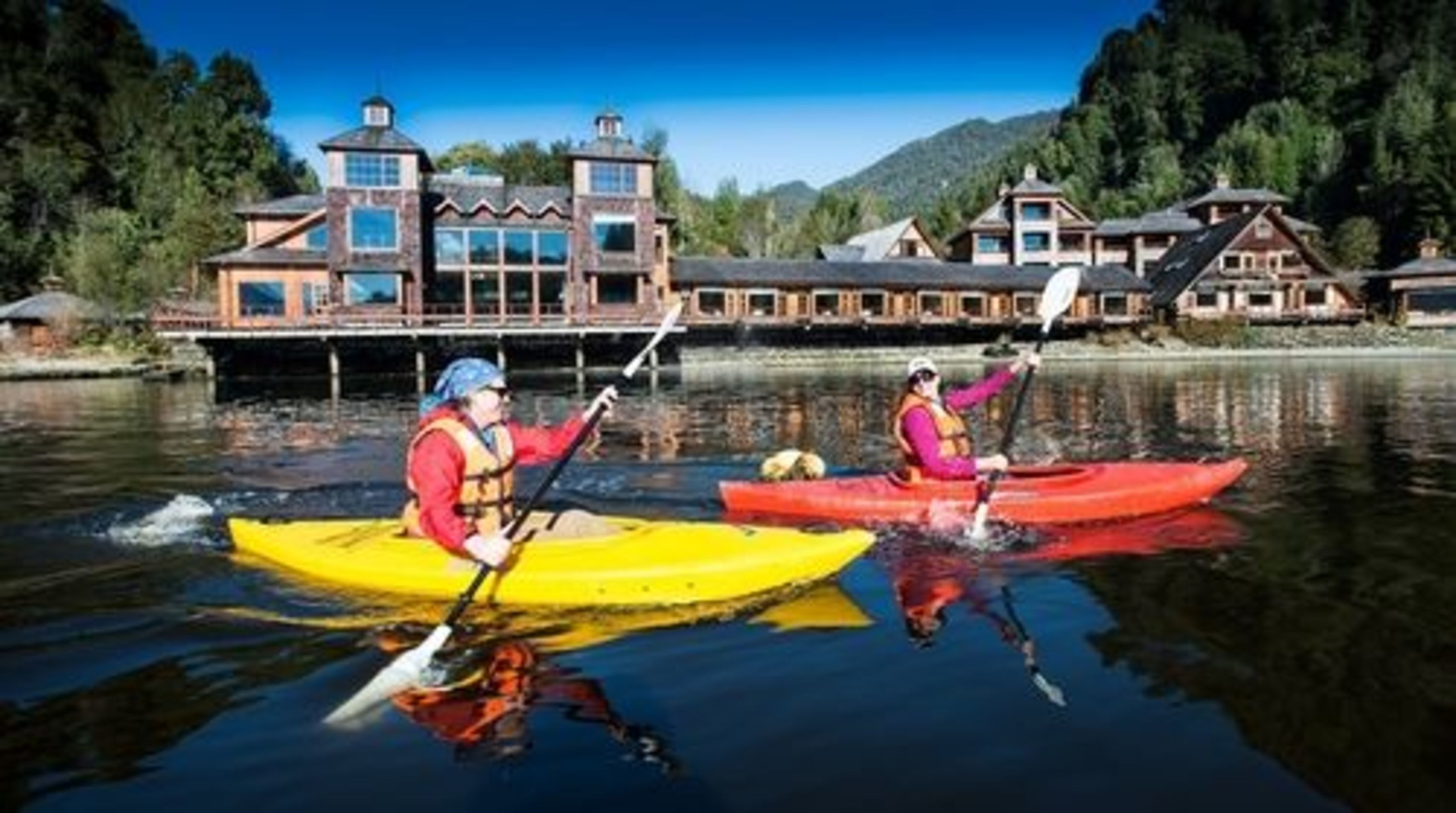 Two people kayaking on a calm river with a lodge in the background.