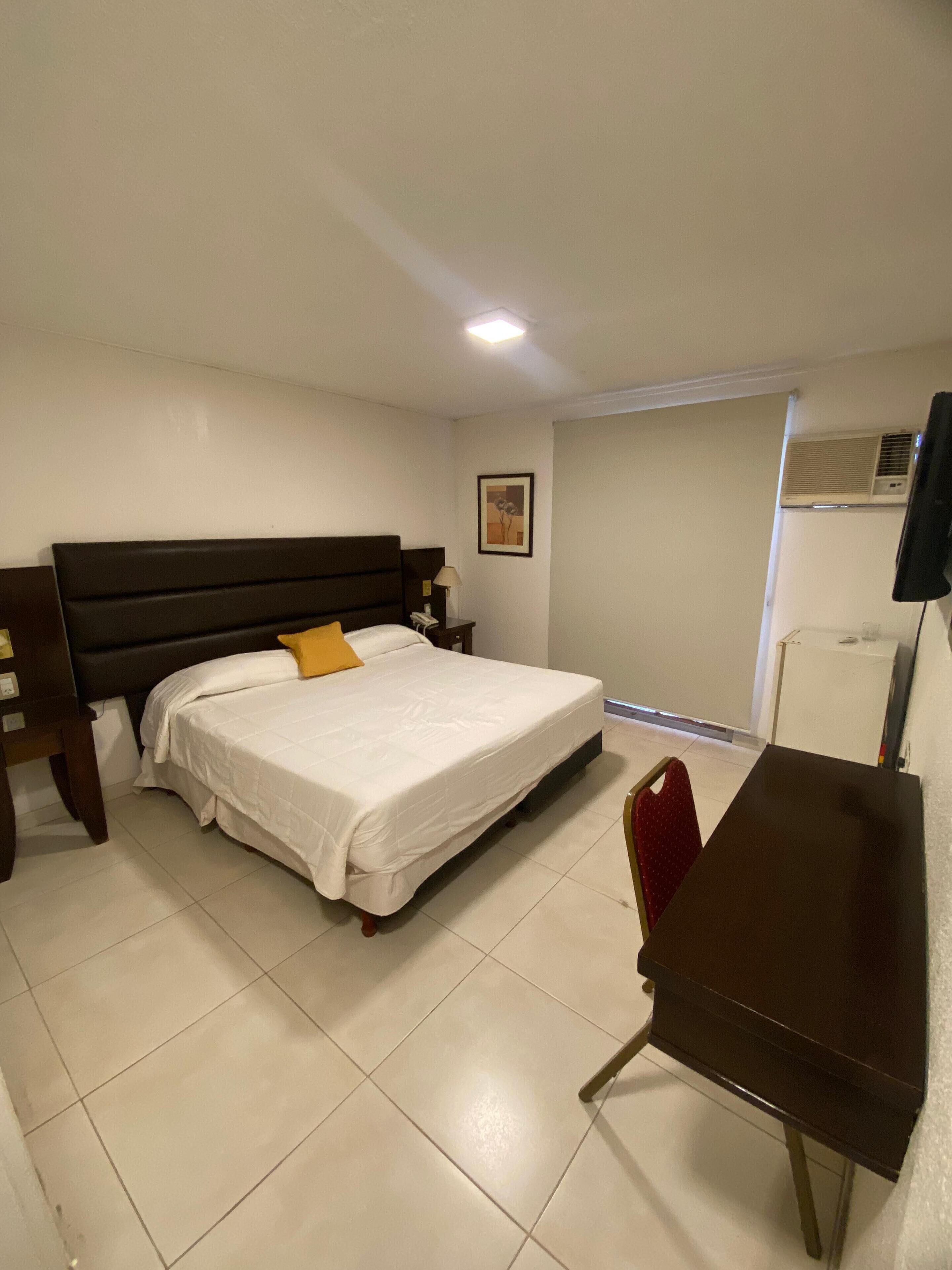 A hotel room with a bed, bedside table, lamp, and a desk with a chair.