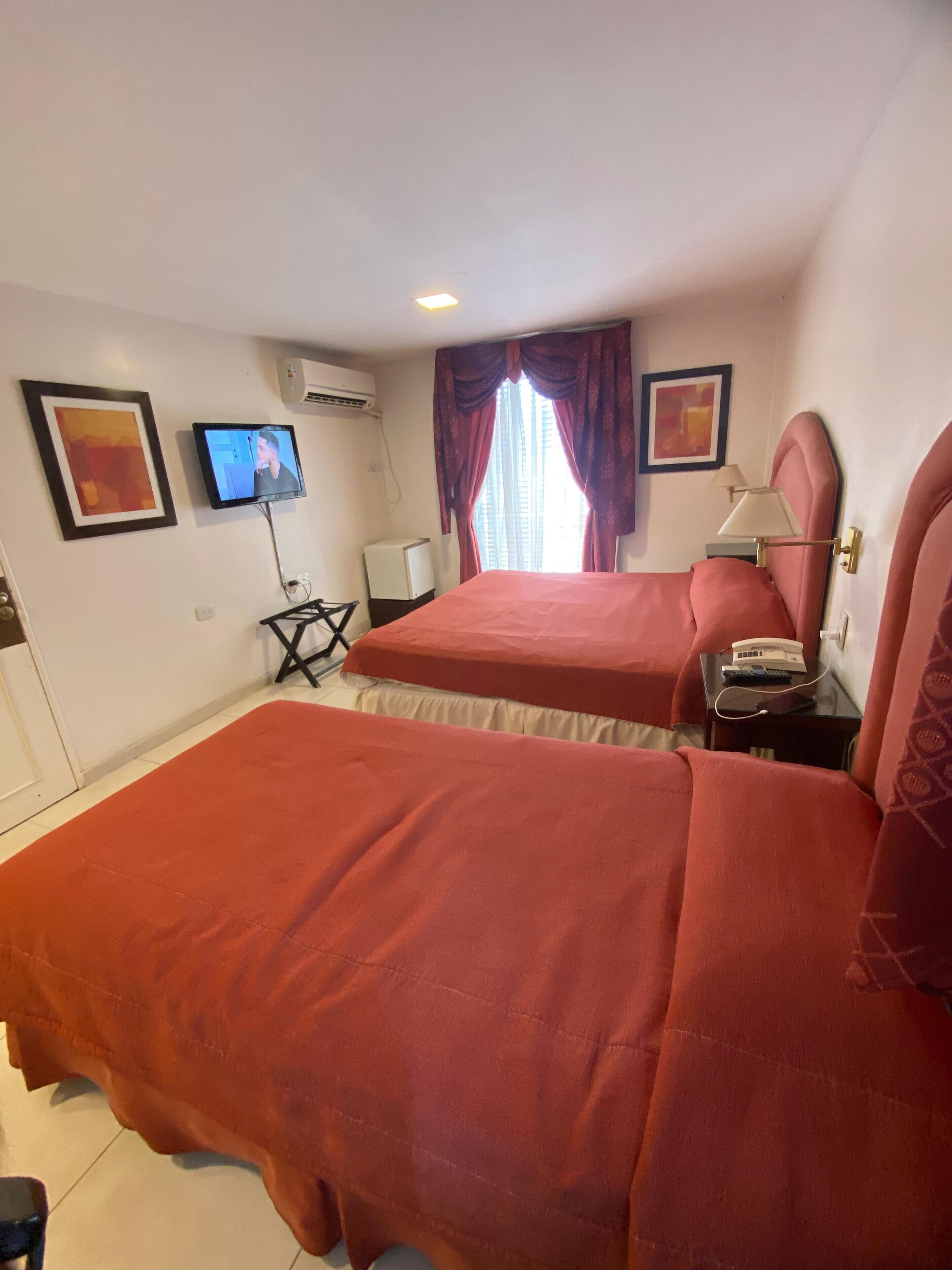 A hotel room with two beds, a TV, an air conditioner, and a small table with a lamp.