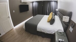 Hotellihuone, jossa on sänky, taulutelevisio, ilmastointilaite, yöpöytä lampun kanssa ja kaukosäädin.