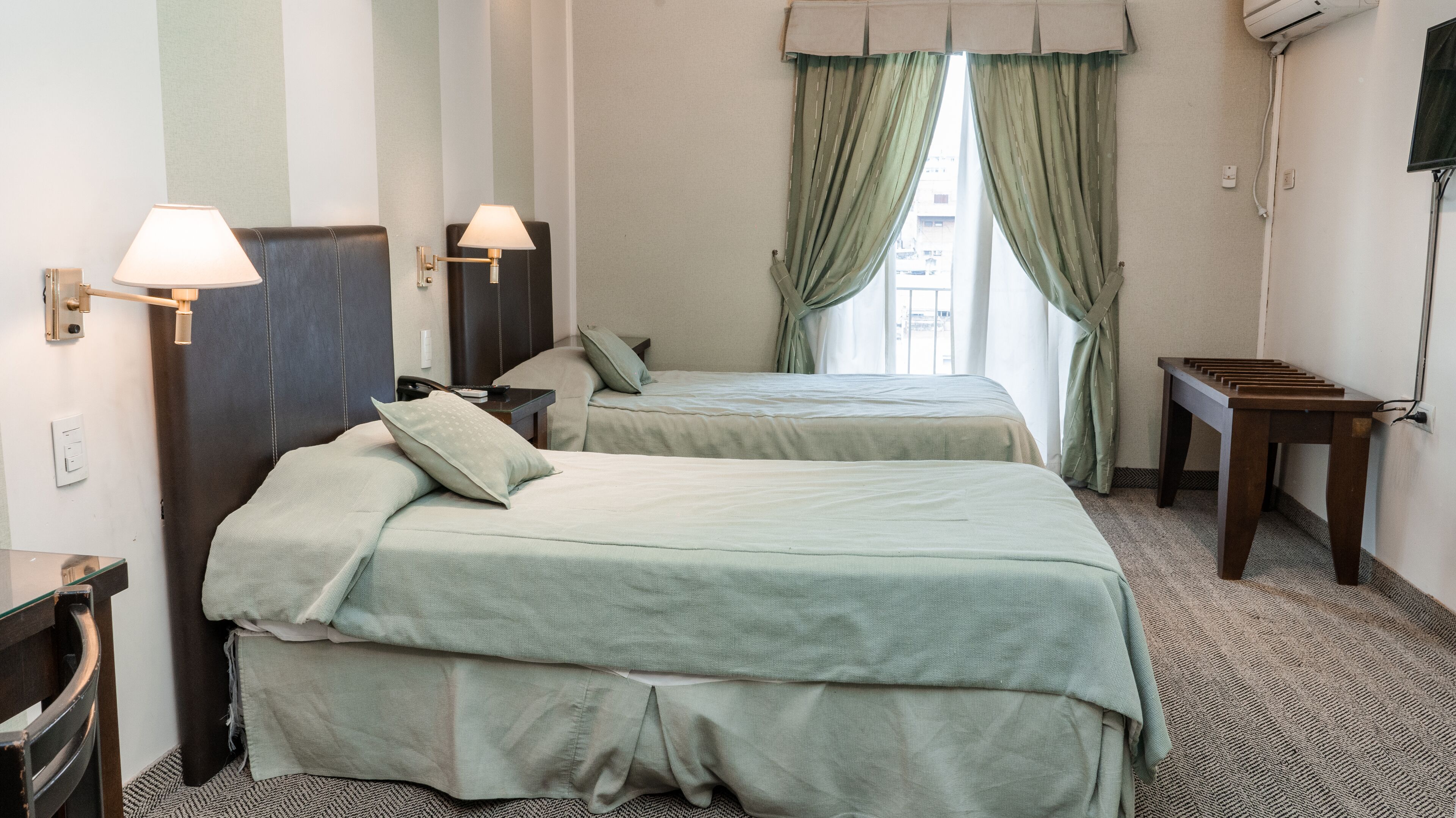 A hotel room with two beds, a desk, a chair, a lamp, and a window with curtains.