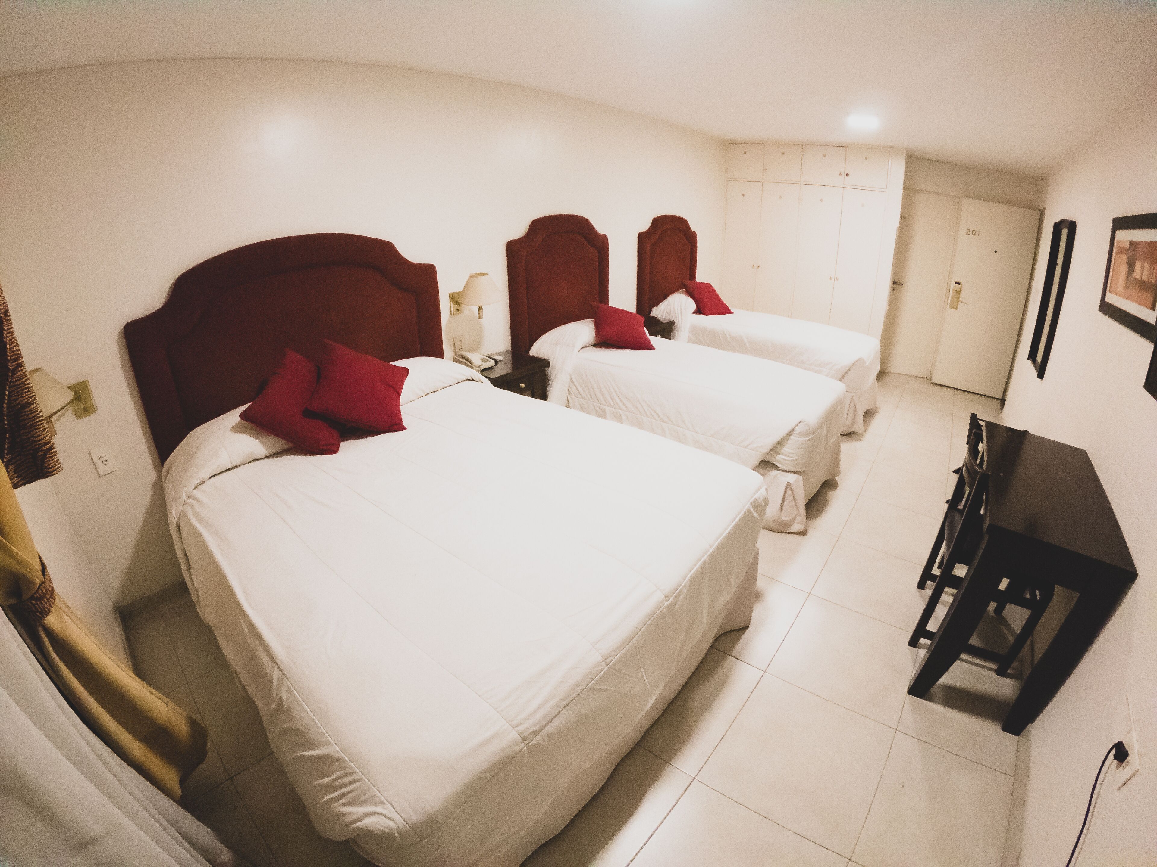 A hotel room with three beds, each with a red headboard and white bedding. There is a desk and a chair in the room.