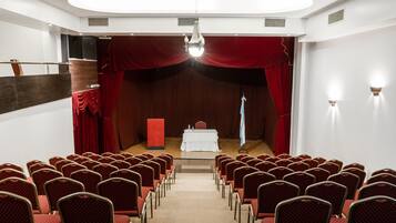 A conference room with rows of red chairs, a stage with a white table, and red curtains.