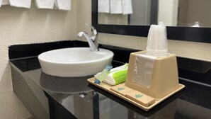 Un lavabo con espejo, un perchero con toallas blancas, y una bandeja con artículos de aseo que incluyen una botella, un paquete verde y una caja beige.