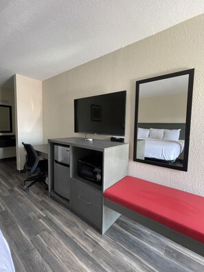 Habitación de hotel con cama, escritorio, computadora, silla, televisor y espejo.