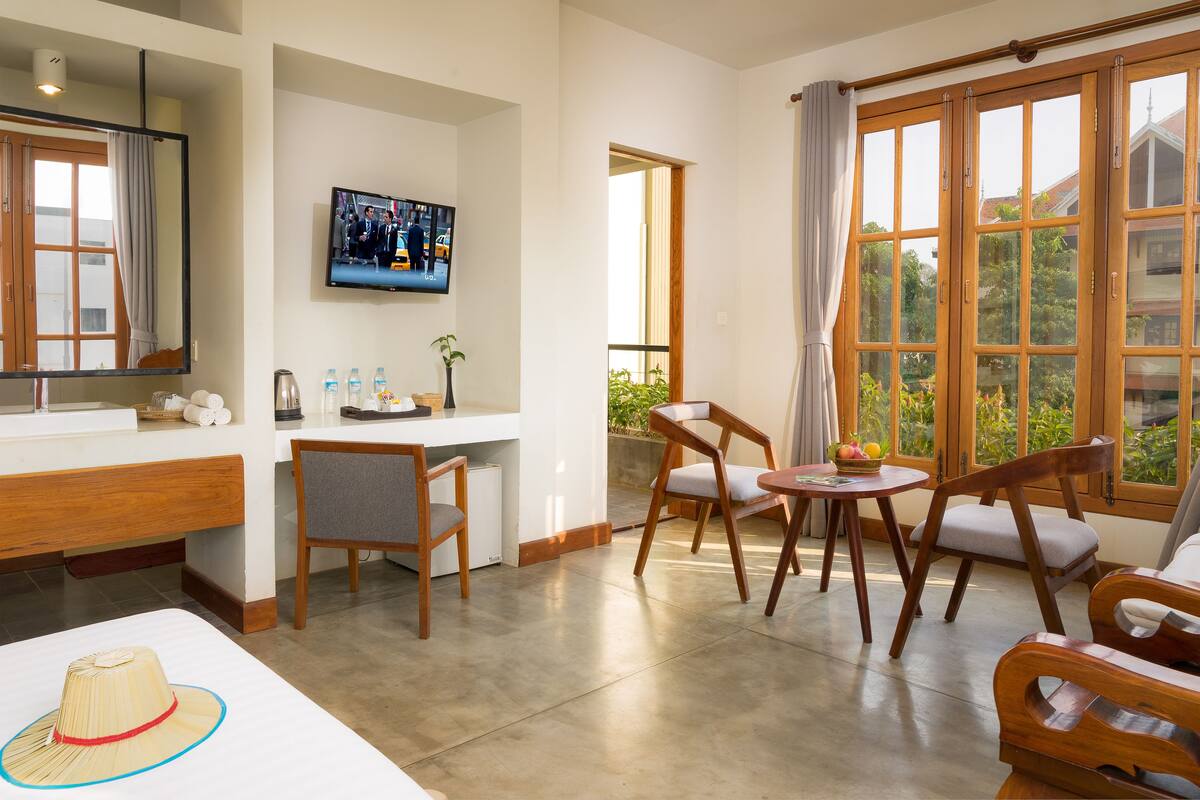 Hotelkamer met een bed, bureau, televisie en balkon met houten deuren en uitzicht op groen.