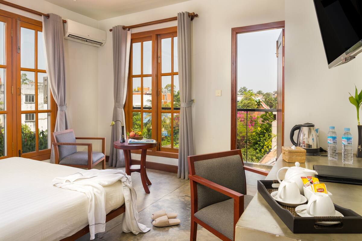 Hotelkamer met een bed, twee stoelen, een kleine tafel met een schaal fruit, een dienblad met kopjes en Lipton thee, een televisie, en een balkon met uitzicht op gebouwen en groen.