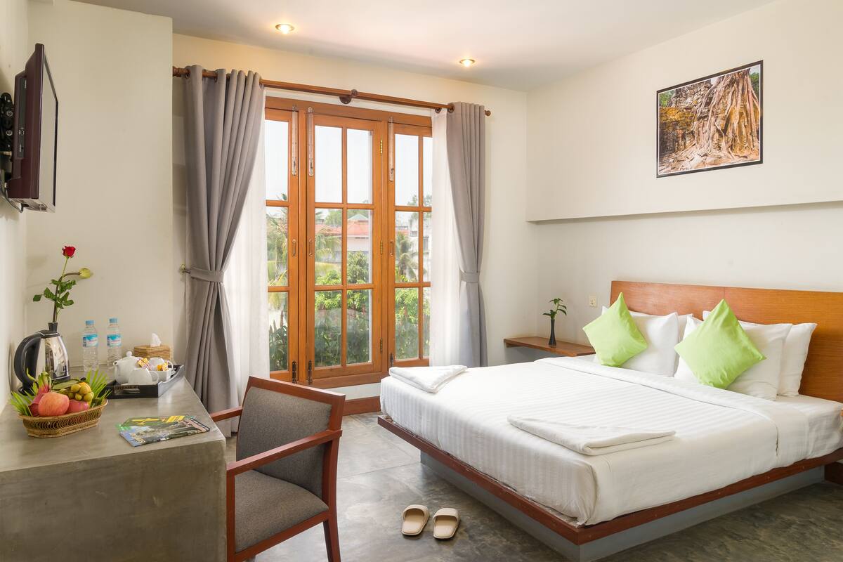 Een hotelkamer met een bed, een bureau met stoel, een raam met gordijnen, een schilderij aan de muur en uitzicht op groen.