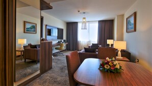 Una habitación de hotel con zona de comedor, televisión, escritorio y un amplio ventanal con cortinas.