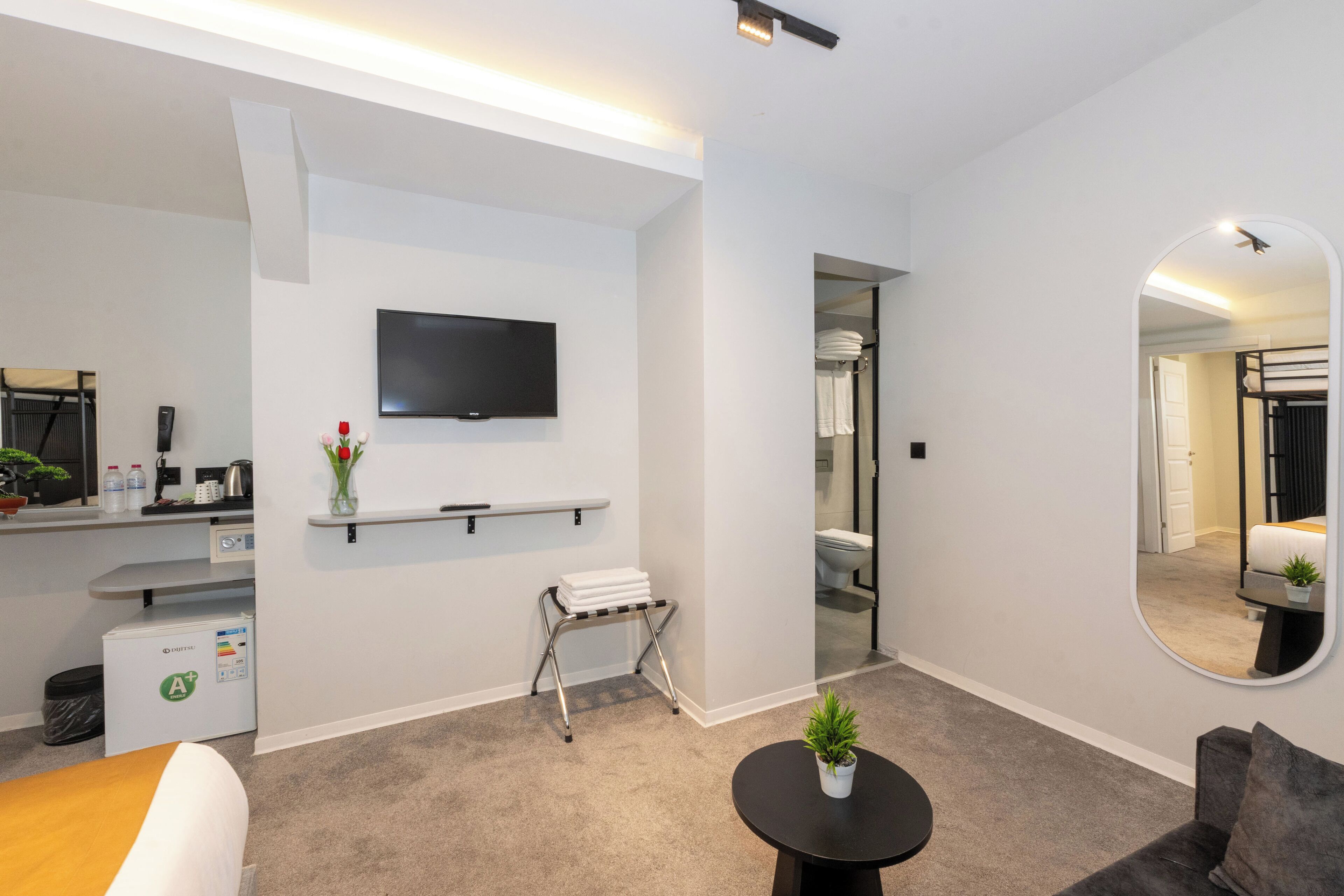 Quarto de hotel moderno com televisão de tela plana, uma pequena mesa com uma planta, sofá, espelho e área de cozinha compacta com micro-ondas e geladeira.