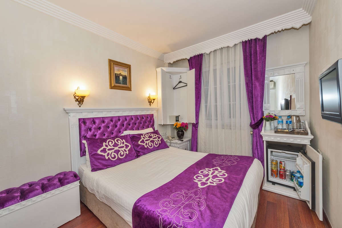 A bedroom with a tufted headboard, a bed with purple bedding, a nightstand with a mirror, and a minibar.