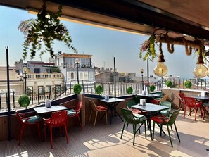 A rooftop terrace with tables and chairs, a view of the city, and hanging plants.