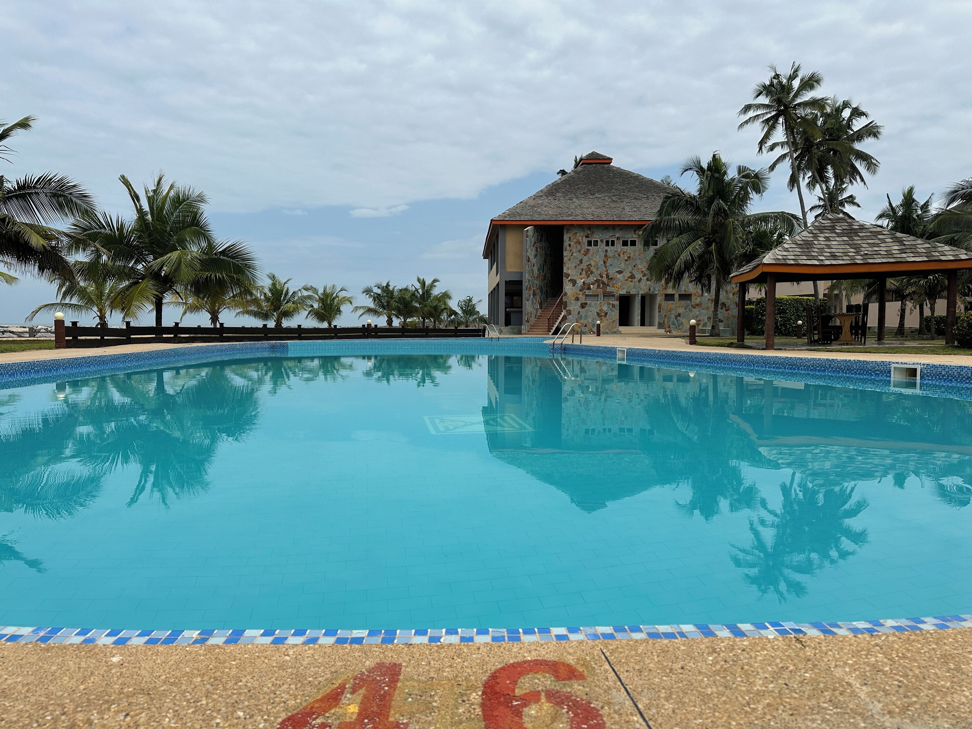 A swimming pool with a tiled edge, surrounded by palm trees and a building with a tiled roof.
