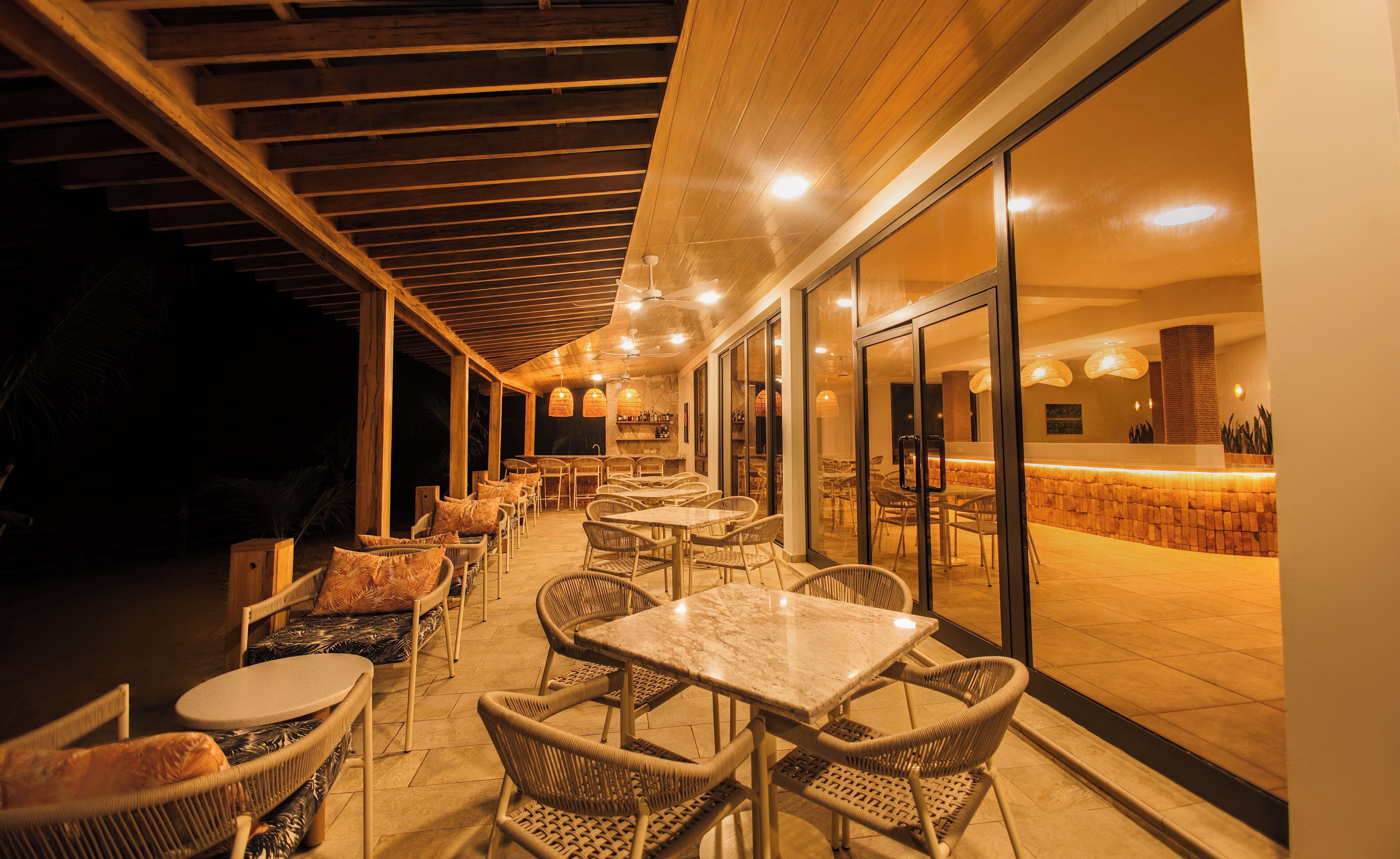Un espace de restauration en plein air avec des chaises et des tables en osier, un coin bar avec un mur en pierre, et un plafond orné de poutres en bois et doté d’un éclairage encastré.