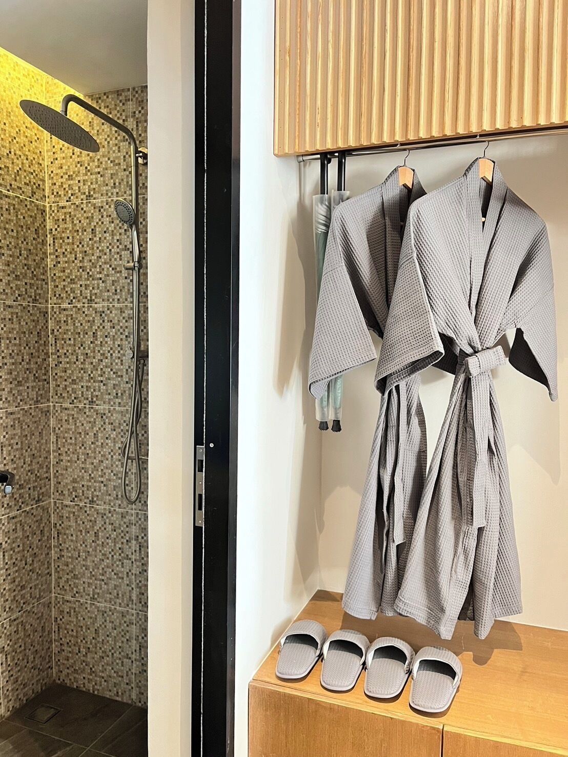 A bathroom with a shower, a wooden vanity with a robe and slippers, and a towel rack.