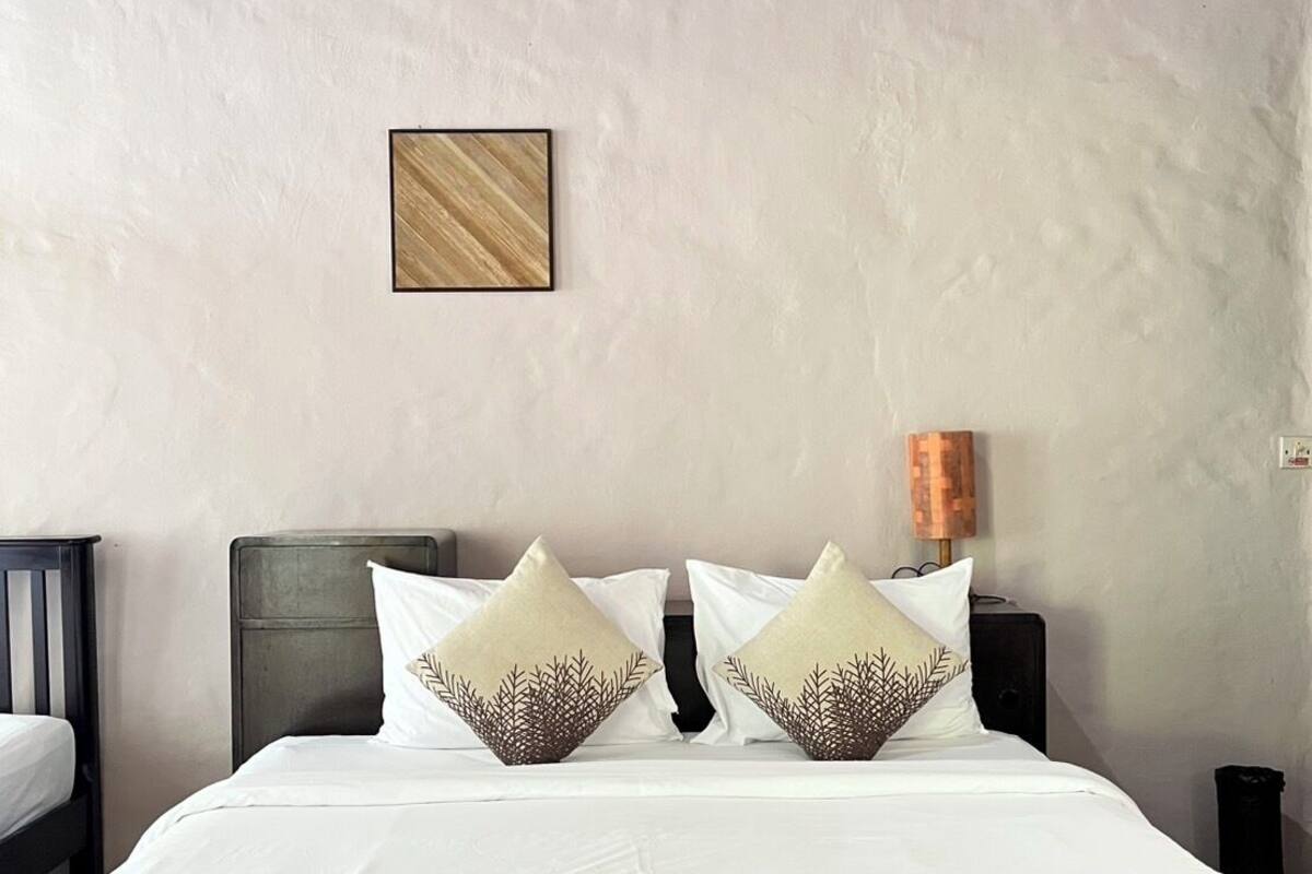 A neatly made bed with white bedding and two decorative pillows, a small framed picture on the wall, and a lamp on the nightstand.