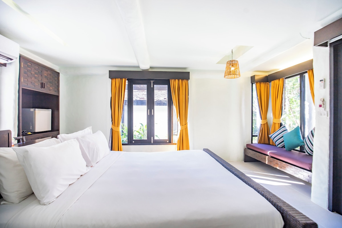 A hotel room with a large bed, a built-in bench with cushions, a ceiling fan, and a view of greenery through the windows.