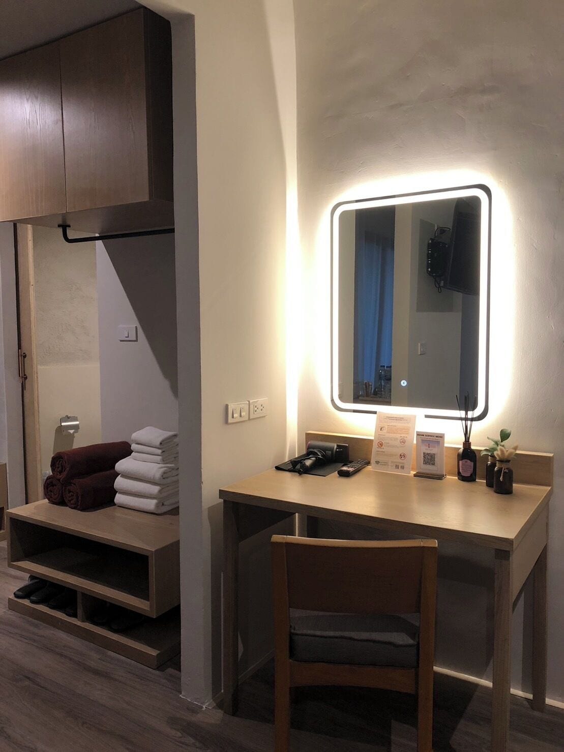 Habitación de hotel con escritorio de madera, una silla, un espejo con iluminación LED, un perchero y un estante con zapatos.
