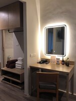 Ett hotellrum med ett skrivbord i trä, en stol, en spegel med LED-belysning, en handduksställning och en hylla med skor.