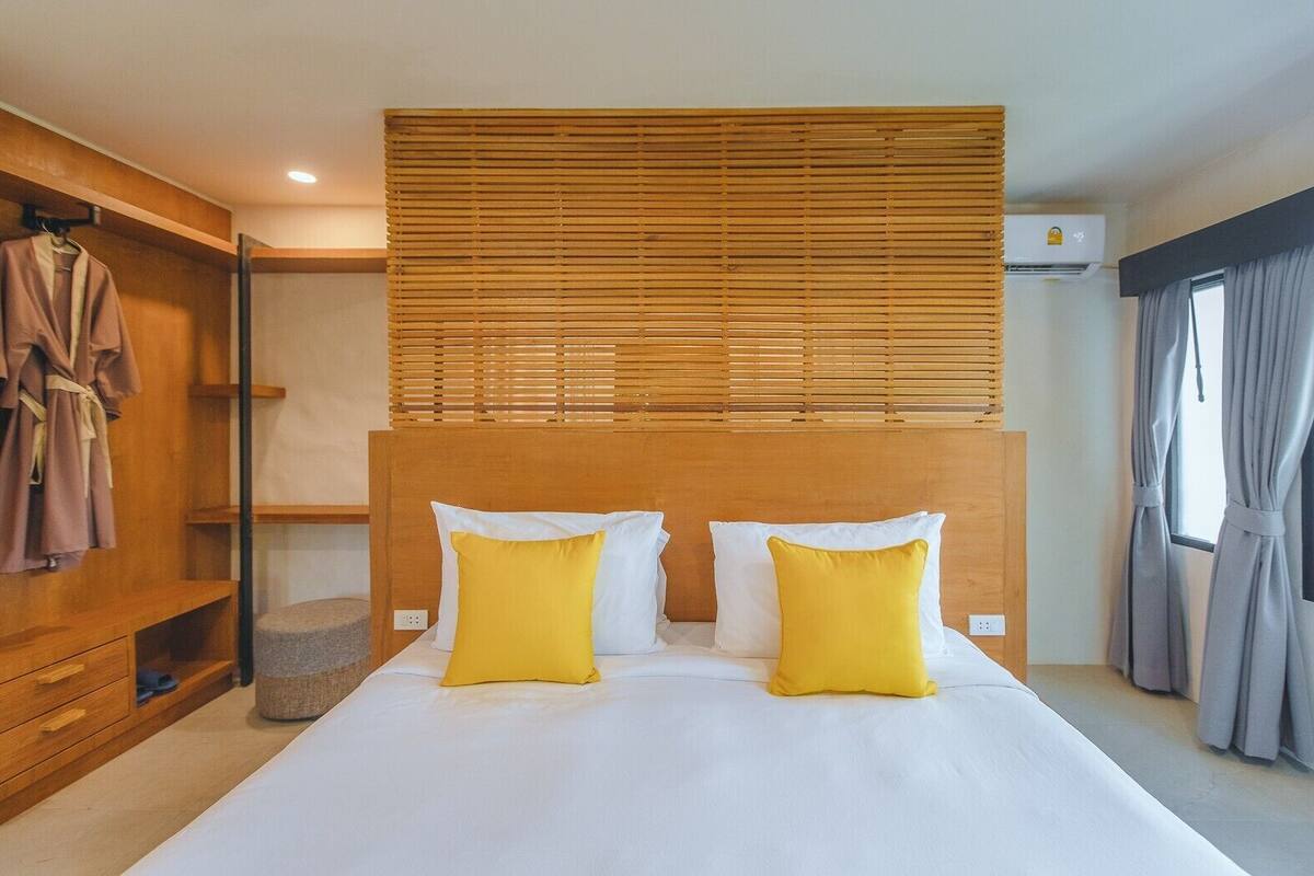 A hotel room with a wooden headboard, a bed with white and yellow pillows, a wooden wardrobe, and a window with curtains.