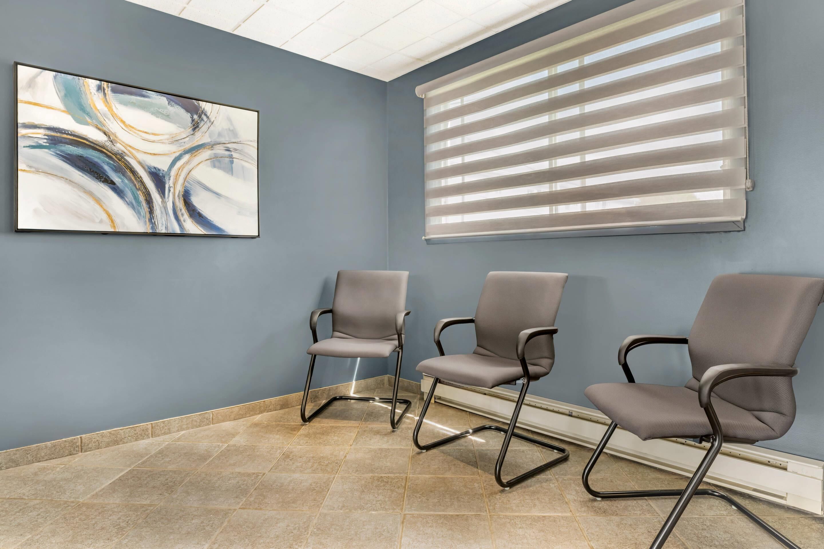 A waiting area with three grey chairs, a blue wall, a large abstract painting, and a window with blinds.